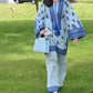 A woman walking on grass wearing a blue traditional two-piece outfit with floral and geometric patterns, including wide-leg pants and a top with bell sleeves.