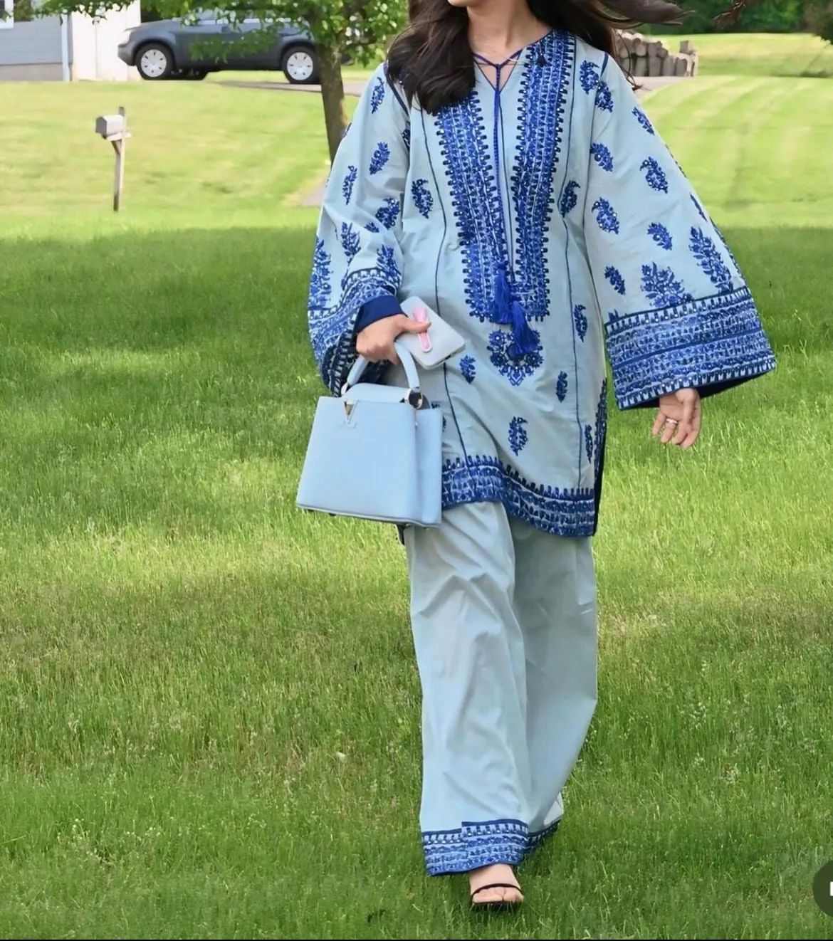 A woman walking on grass wearing a blue traditional two-piece outfit with floral and geometric patterns, including wide-leg pants and a top with bell sleeves.